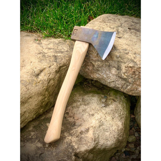A natural wood-handled Council Tool Co. Hudson Bay Camp Axe rests on stacked rocks with green grass in the background.