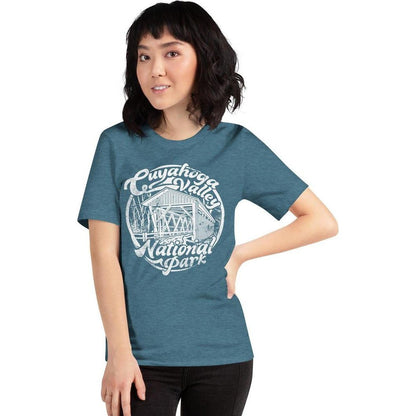 A woman wearing a Heather Deep Teal Cuyahoga Valley Clothing Company CVNP Covered Bridge T - Shirt stands against a white background.