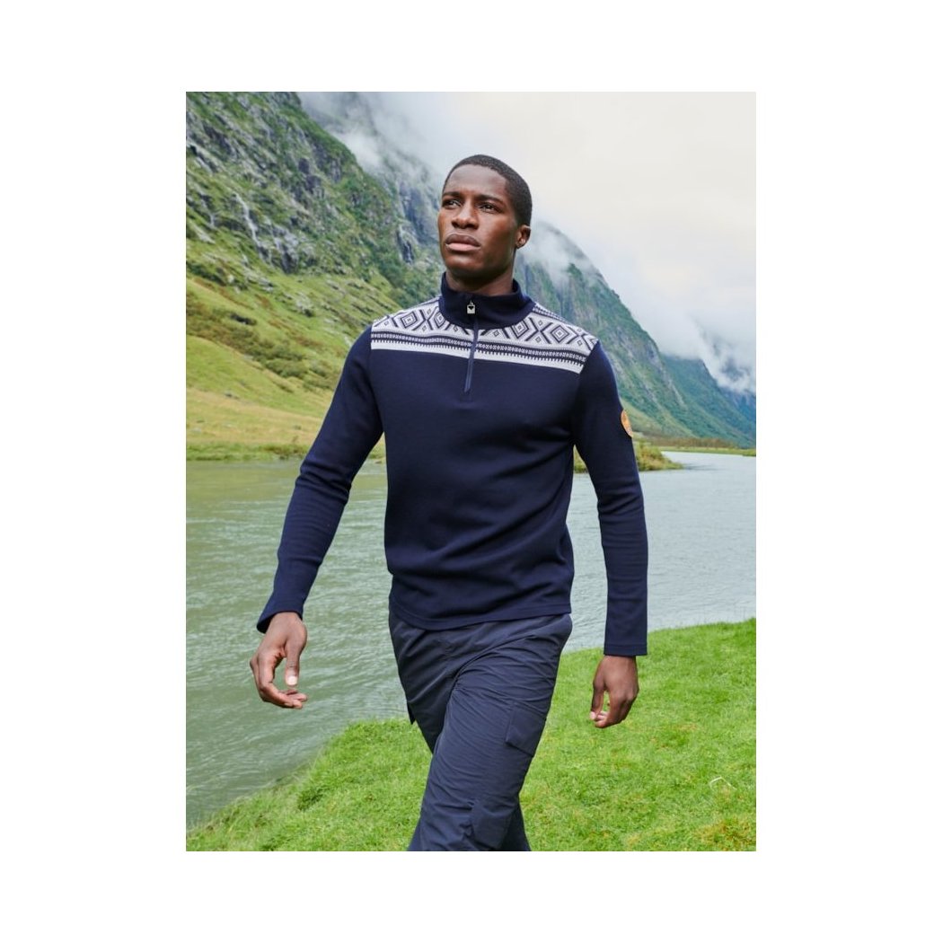 A man wearing the Dale Of Norway Men's Cortina Basic Sweater-SAMPLE in Navy walks near a lake with mountains in the background.