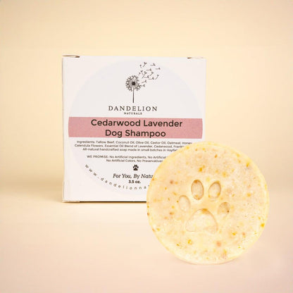 A front view of the Dandelion Naturals Cedarwood Lavender Dog Shampoo Bar with a paw print on it in front of its box against a light background.