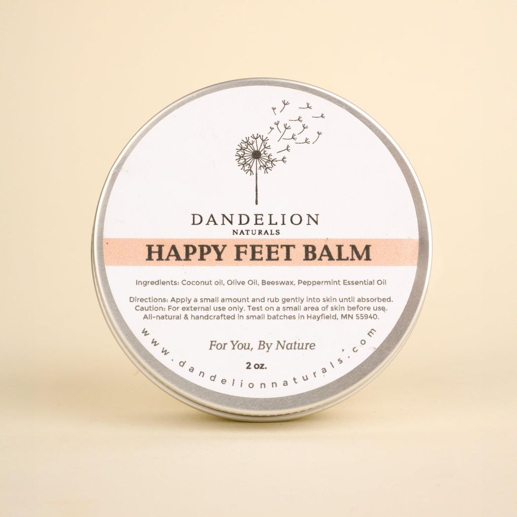 A round tin of Dandelion Naturals Happy Feet Balm sits centered against a plain beige background.