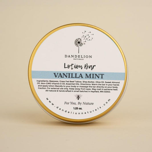 A front view of the Dandelion Naturals Vanilla Mint Tallow Lotion Bar in a round tin with a gold rim against a plain beige background.