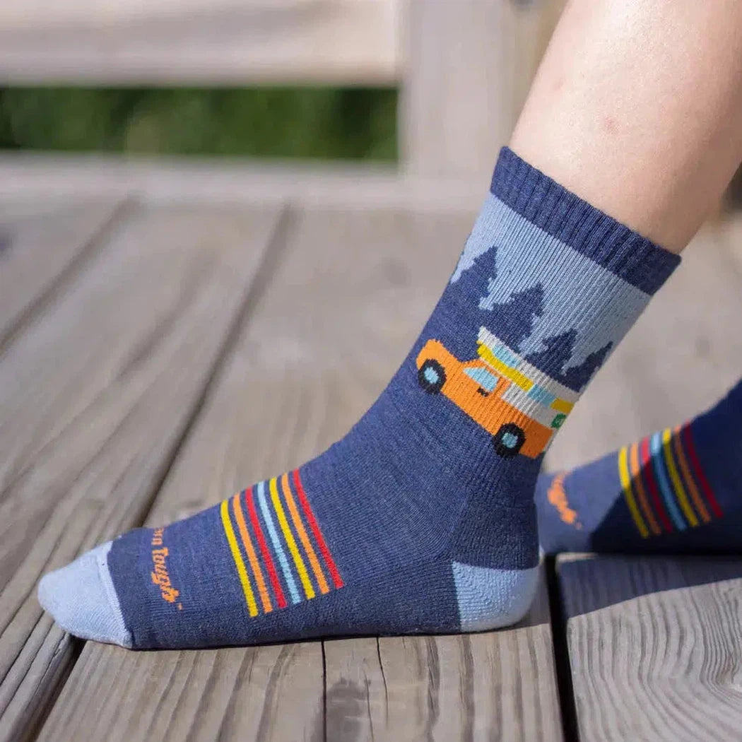 A child’s leg in a Denim Darn Tough Kid's Wild Micro Crew Lightweight with Cushion sock sits on a wooden deck with a forest background.