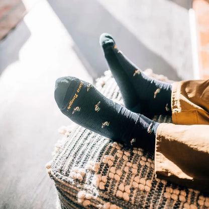 A close-up of a man’s feet wearing Darn Tough Men's Duck Duck Moose Crew Lightweight with Cushion socks in Eclipse, resting on a woven ottoman with a light-colored floor in the background.