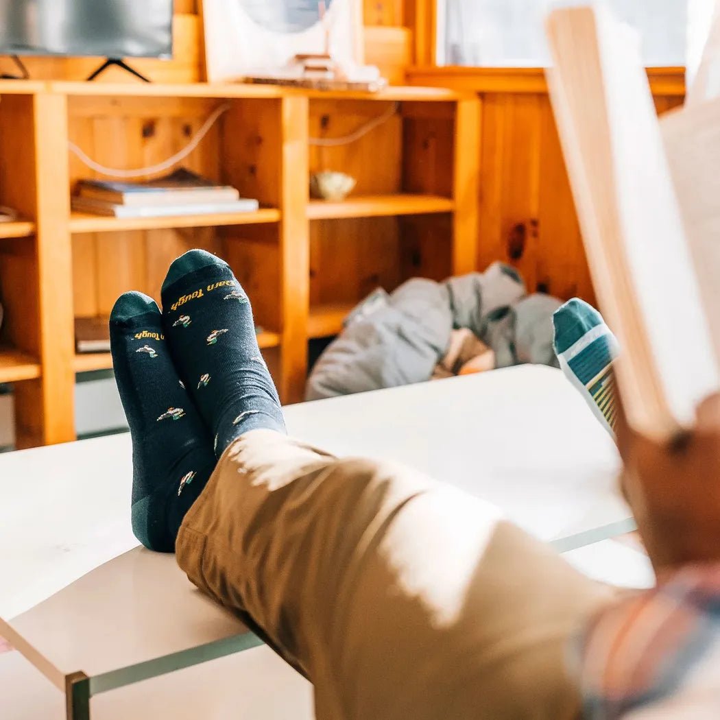 A man relaxes with his feet up on a table wearing Darn Tough Men's Duck Duck Moose Crew Lightweight with Cushion socks in Eclipse, reading a book in a cozy wooden room.