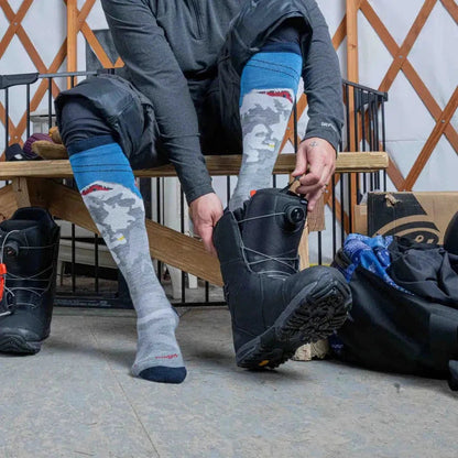 A man puts on Darn Tough Men's Heady Yeti OTC Midweight with Cushion socks in Gray while sitting on a wooden bench.