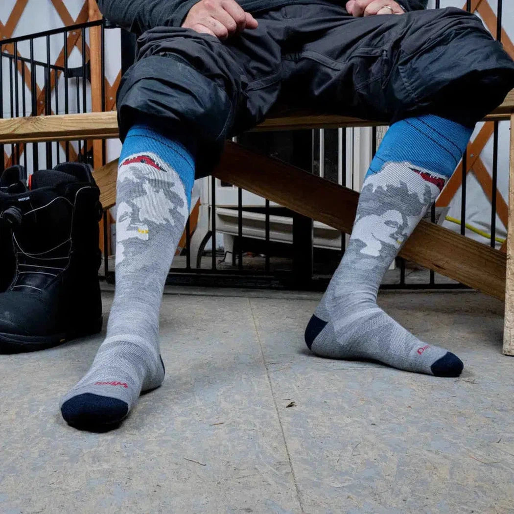 A man wearing Darn Tough Men's Heady Yeti OTC Midweight with Cushion gray socks sits on a wooden bench with black boots nearby.