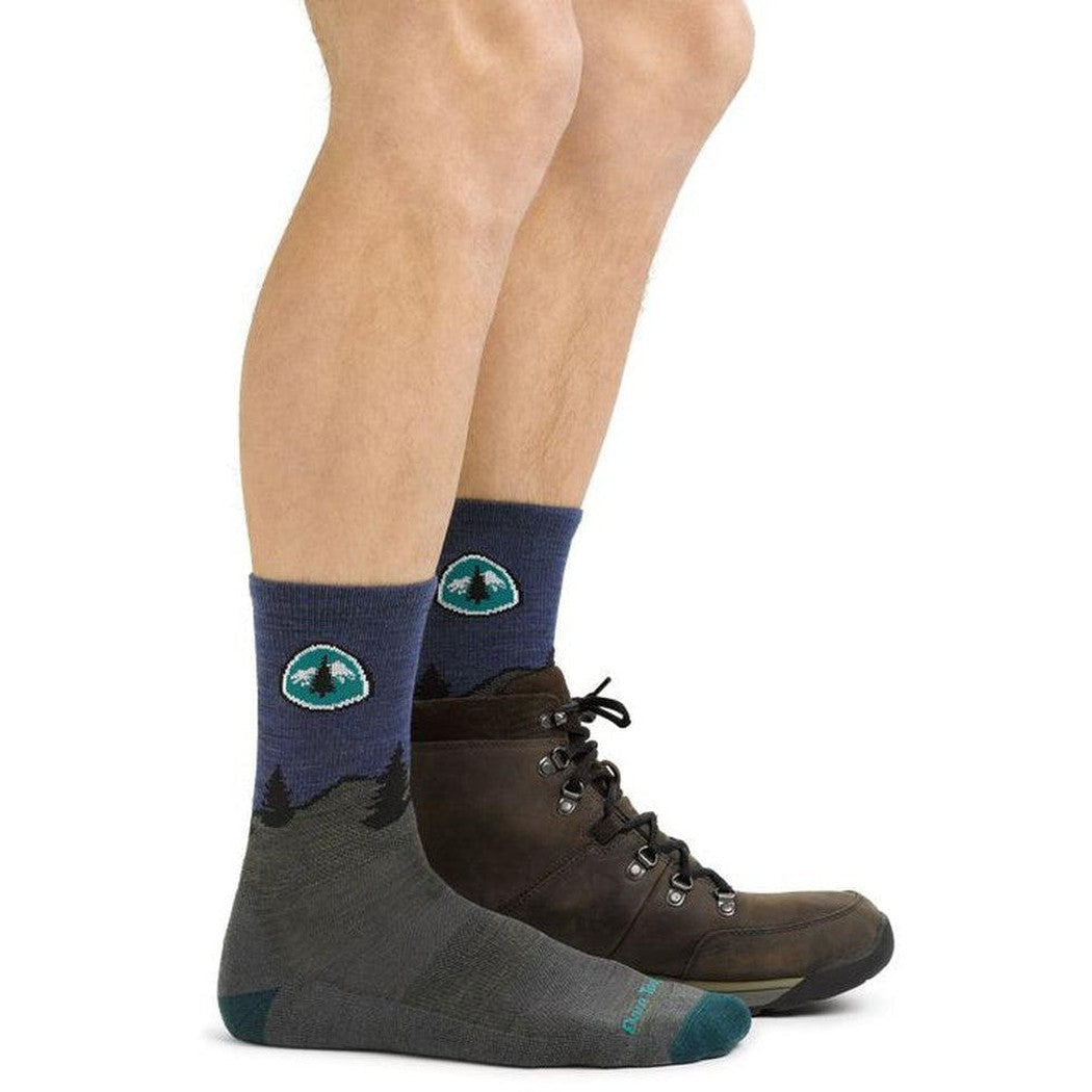 Side view of the Darn Tough Men's PCT Micro Crew Lightweight with Cushion socks in Forest, showing the mountain graphic on the cuff and a model wearing them with brown hiking boots against a white
