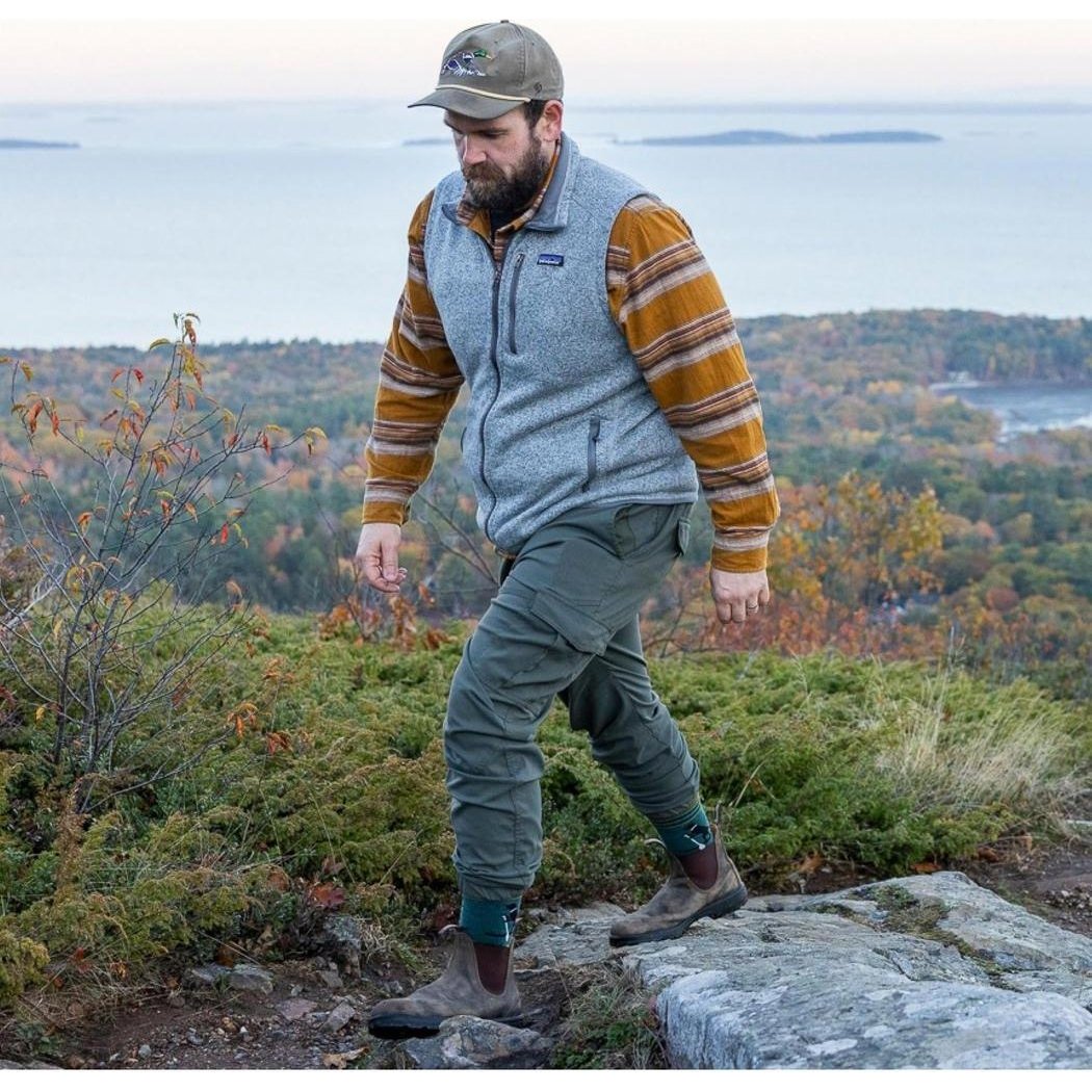 A man hikes on a rocky trail wearing the Darn Tough Men's Willoughby Micro Crew Lightweight Hiking Sock in Dog Pond, with a scenic forest and water view behind him.