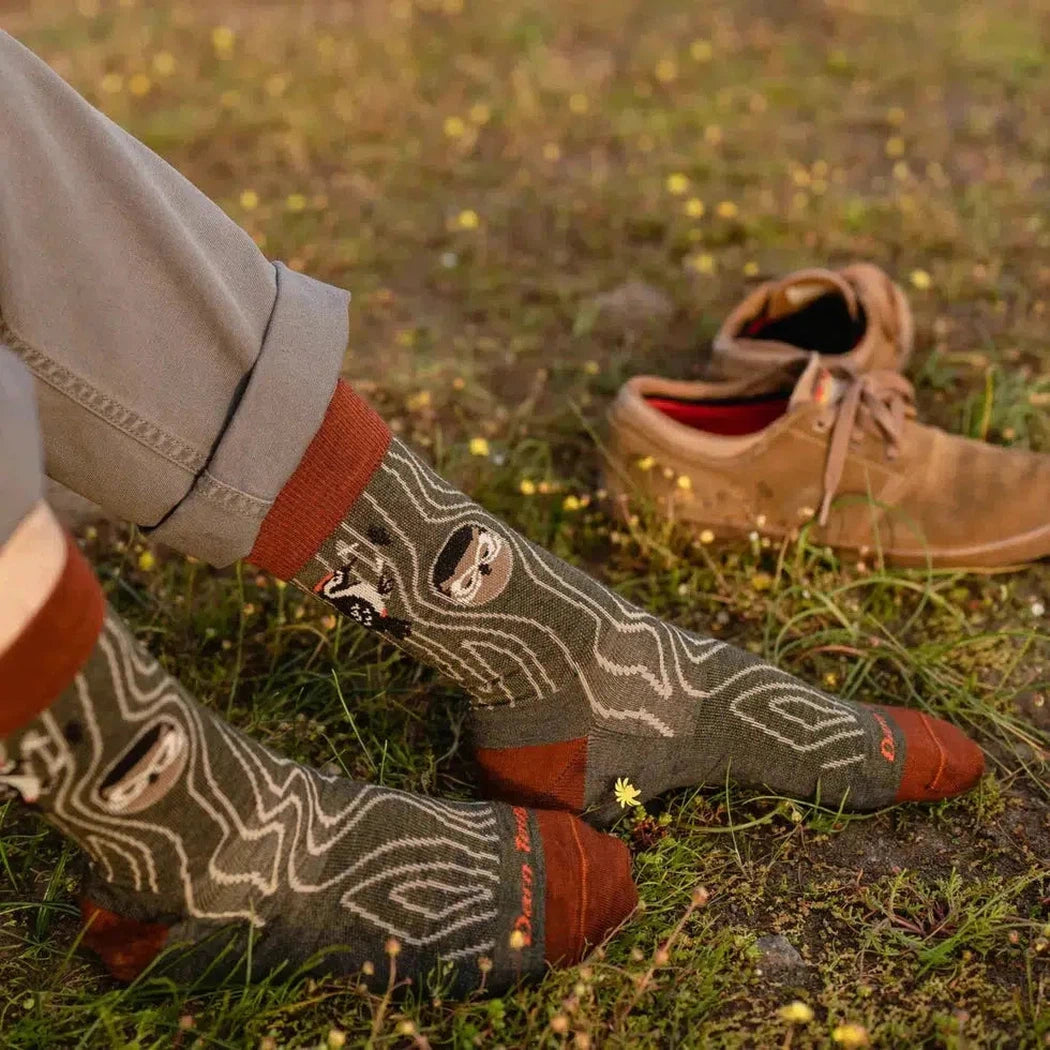 A person wearing Darn Tough Men's Woody Crew Lightweight Forest socks sits on grass with brown shoes nearby.