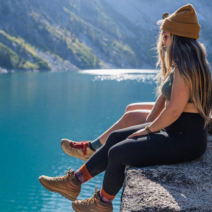 A woman sits by a lake wearing Earth-colored Darn Tough Northwoods Micro Crew Midweight with Cushion socks and tan hiking boots.