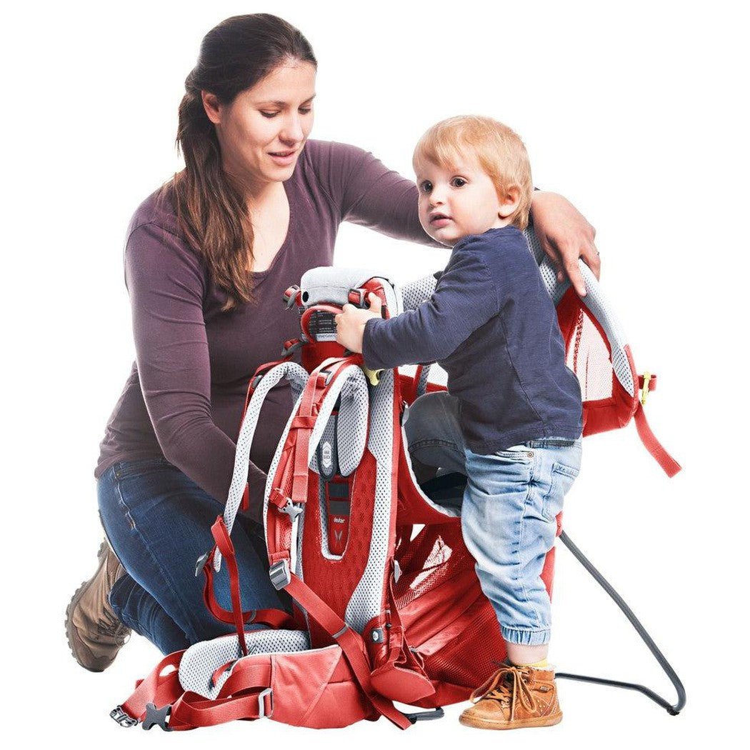 A woman kneels next to a toddler standing in the Deuter Kid Comfort Active SL child carrier in Currant, showing the carrier's straps and padded seat against a white background.