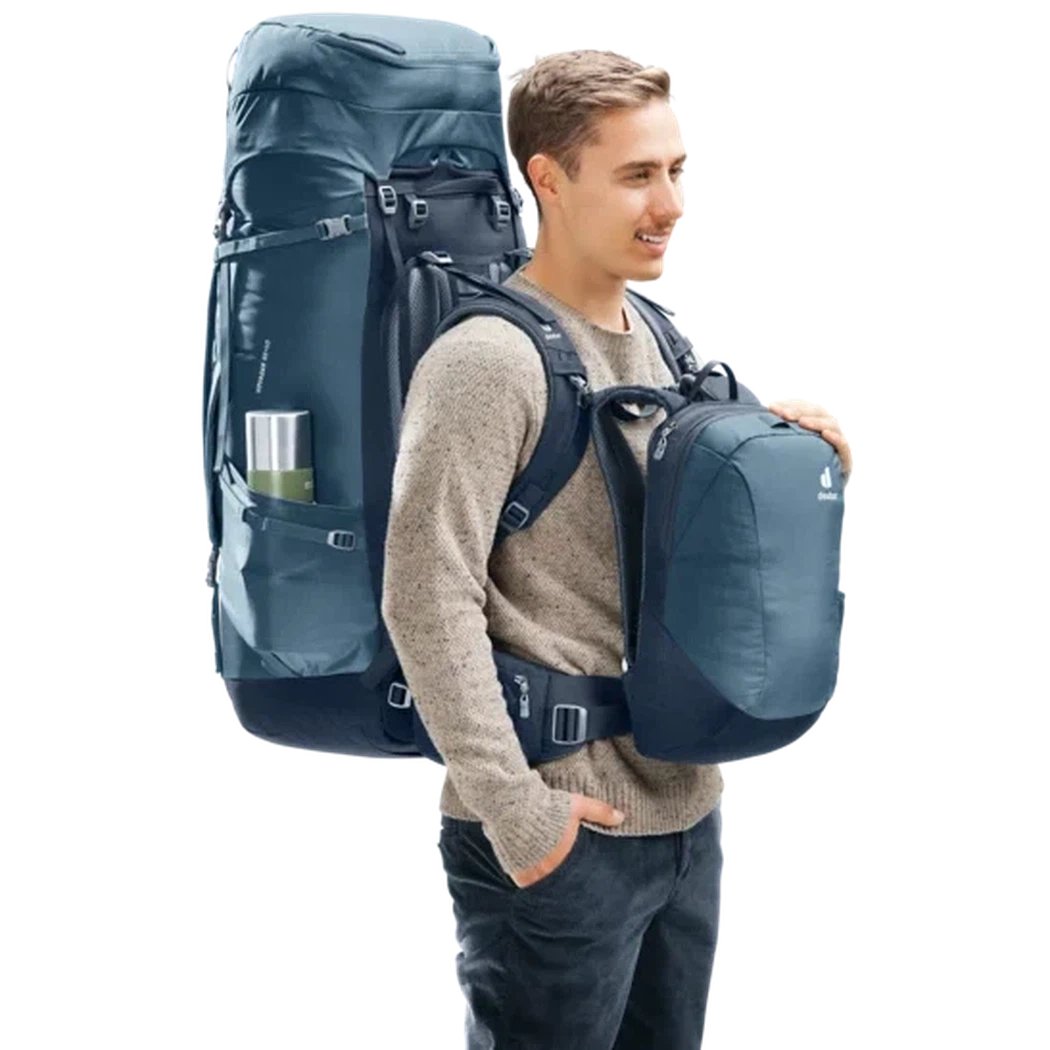 A man wears the Deuter Voyager 65+10 backpack in Atlantic Ink with a smaller bag attached, against a white background.
