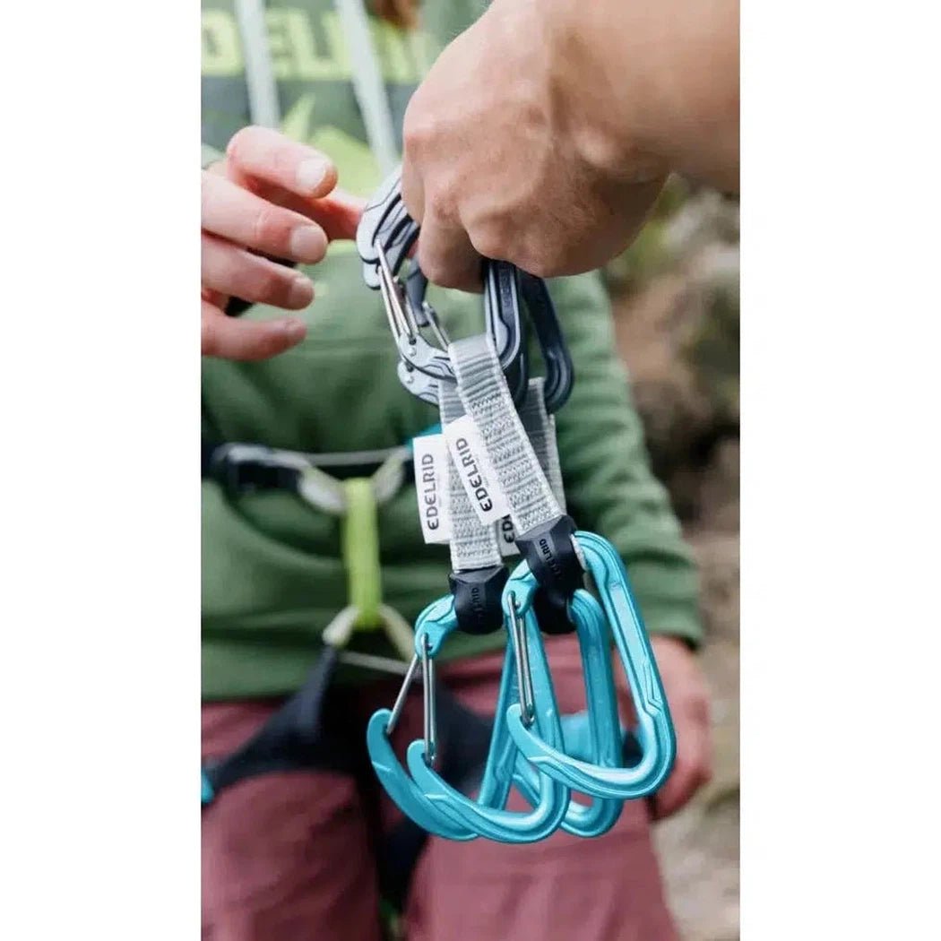A hand holds the Edelrid Pure Wire Set Sixpack, 10cm, with its slate and icemint carabiners visible against a model's green shirt and outdoor background.