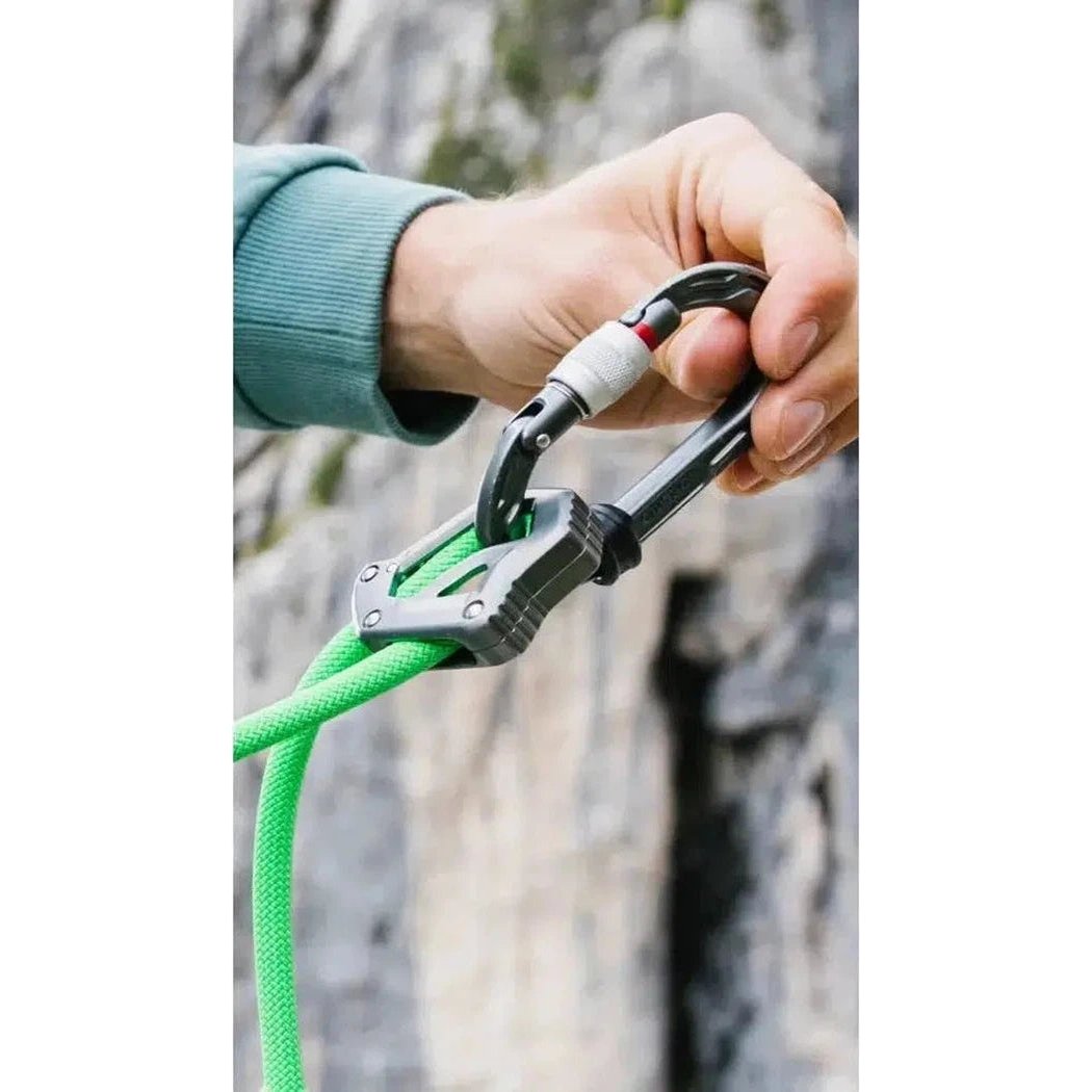 A hand attaches the Edelrid Switch Adjust 100cm carabiner to a Neon Green climbing rope against a rocky background.