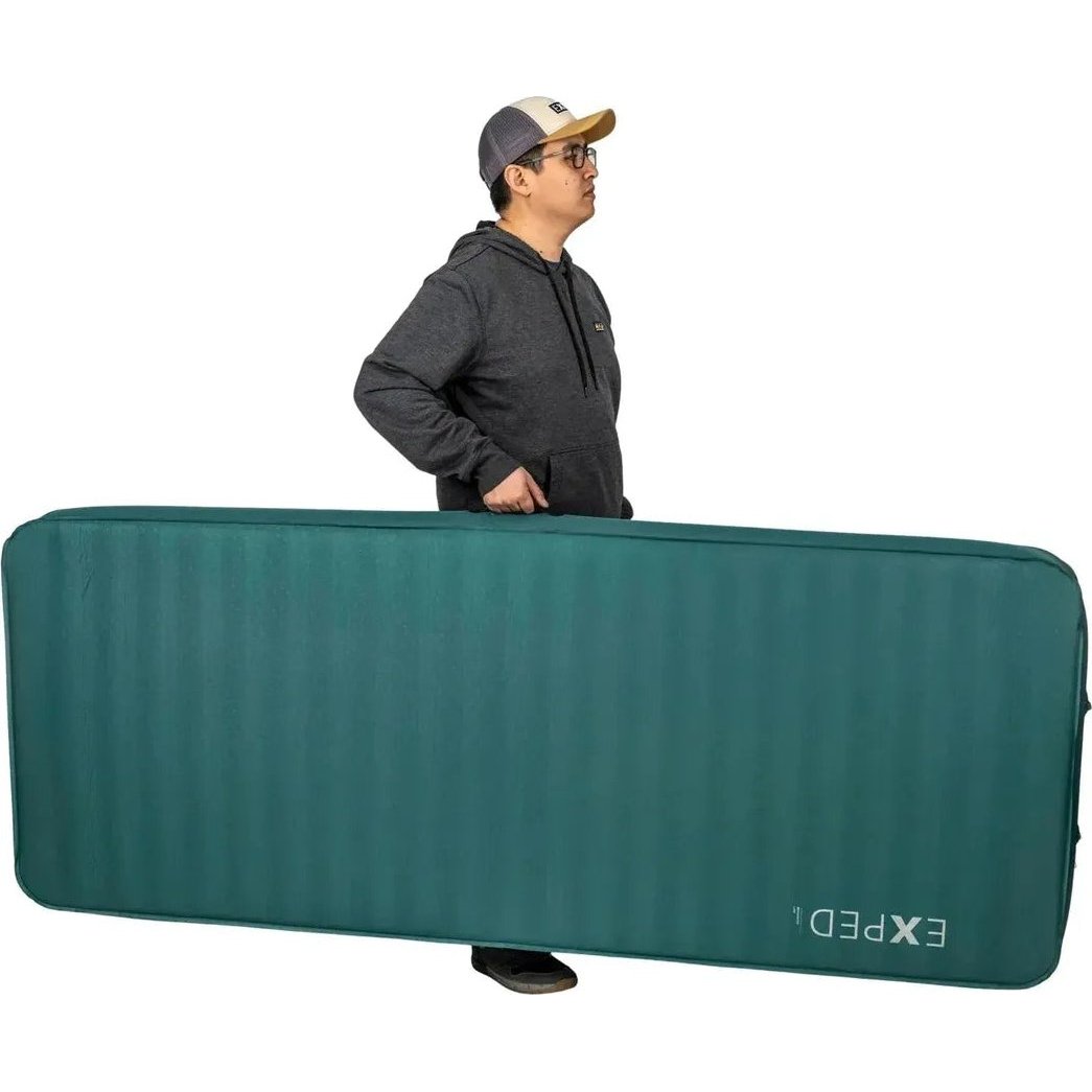 A man in a charcoal hoodie and cap carries the large teal Exped MegaMat against a white background.