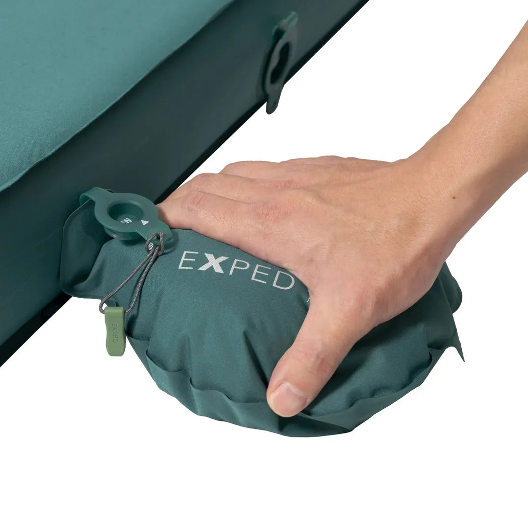 A hand presses the green Exped MegaMat against a teal camping pad, showing its valve and logo.
