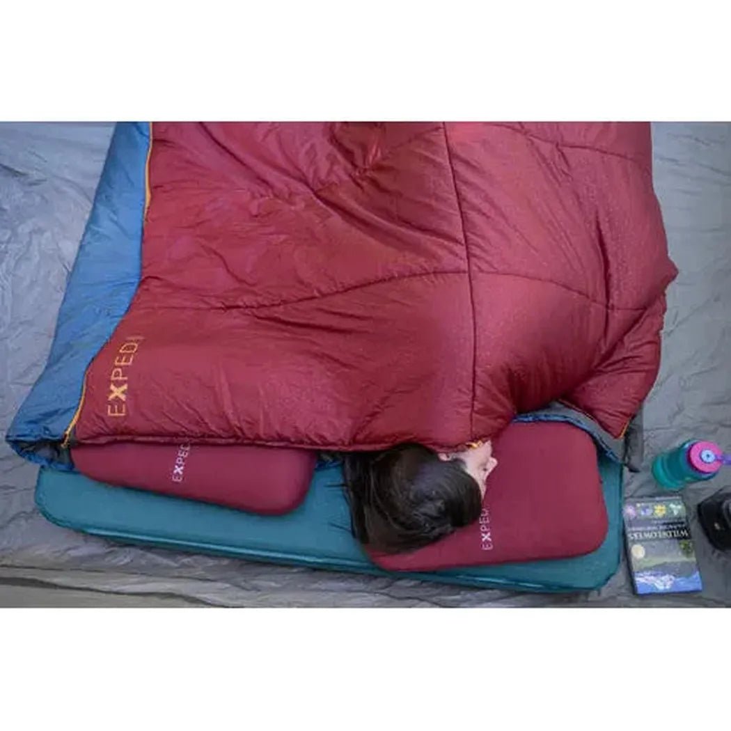 A top-down view of a woman sleeping on a burgundy Exped MegaPillow and matching sleeping pad, with a red sleeping bag and camping gear nearby.