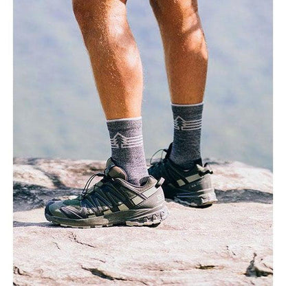 A person wearing FITS Light Hiker (Gifford) Crew socks and shoes stands on a rocky surface with a mountain background.