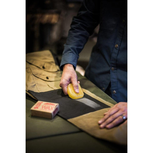 A person applies yellow wax to a dark fabric piece on a table, with a "Fjallraven Greenland Wax" box nearby.