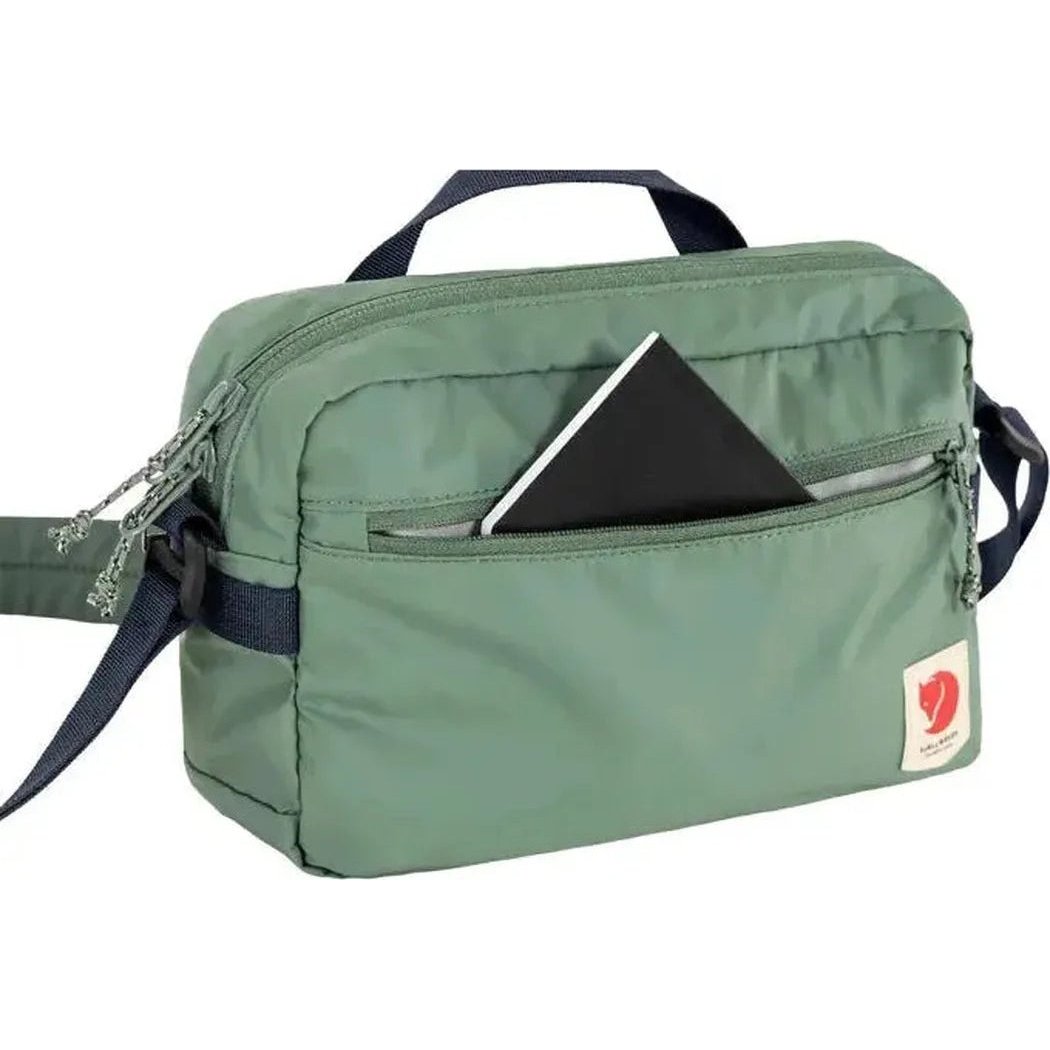 A front view of the Fjallraven High Coast Crossbody in Black with a zippered front pocket holding a black notebook, against a white background.