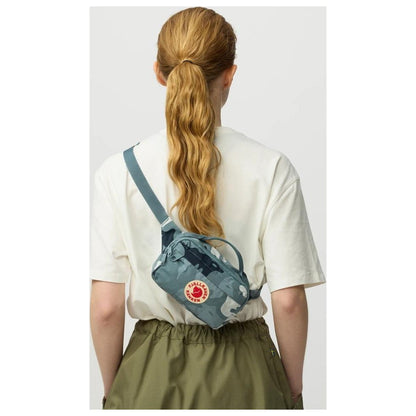 Back view of the Fjallraven Kanken Graphics Hip Pack in Chalk Rose-Hidden Animals on a model wearing a white tee and olive skirt against a light gray background.