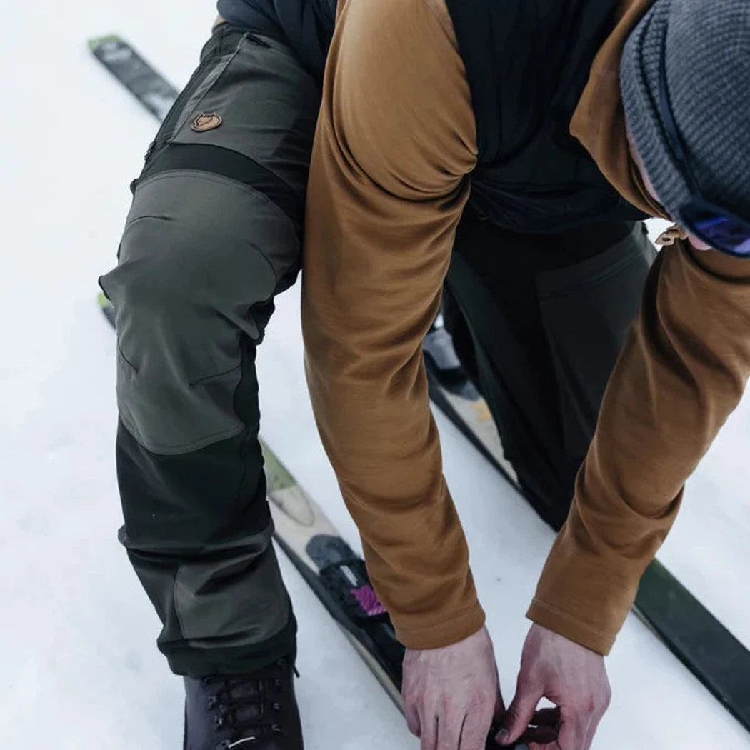 A man in a brown long-sleeve and black Fjallraven Men's Expedition X-Latt Vest adjusts ski boots on a snowy slope.