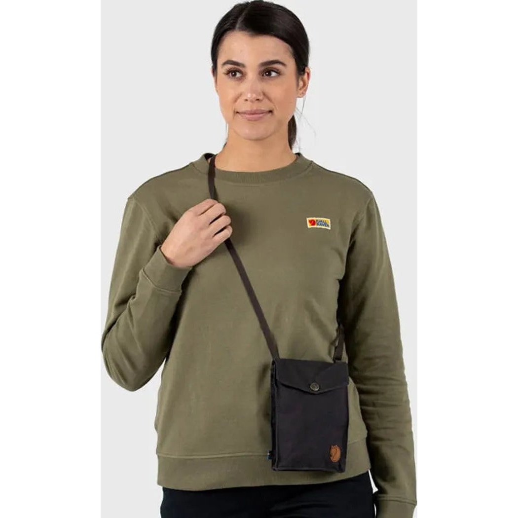A woman wears the Fjallraven Pocket bag in black over her olive sweatshirt, with a white background.