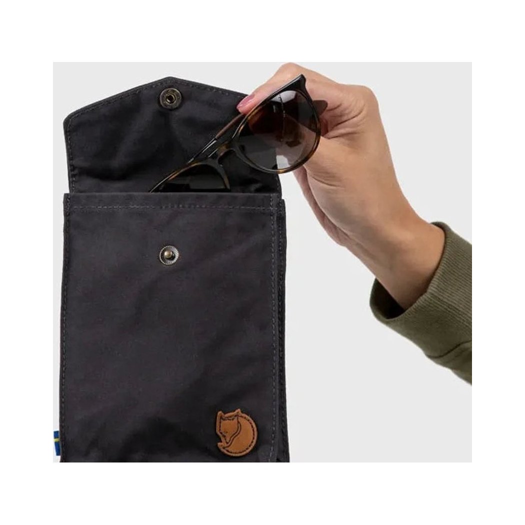 A hand puts sunglasses into the Fjallraven Pocket, a black bag with a fox logo, against a light background.