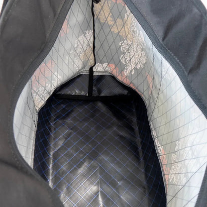 The interior of the Flowfold Jkp X Flowfold | Mammoth Zipper Tote - 29L Tote Bag shows a black and blue checkered bottom and a patterned lining with a zipper pull visible.