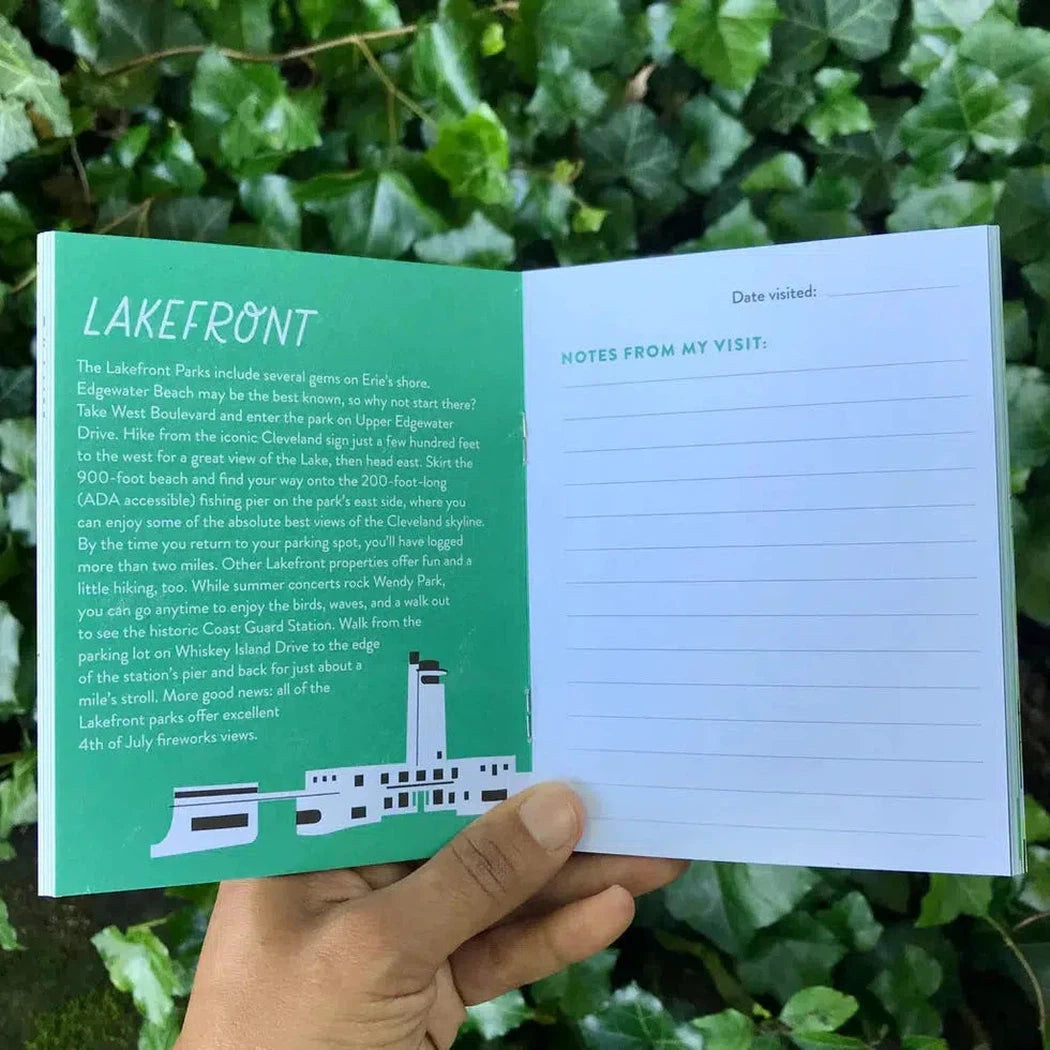 A hand holds the Free Period Press Cleveland Metroparks Mini-Guide open to a green "Lakefront" page with a white "Notes from my visit" page, against a background of green leaves.