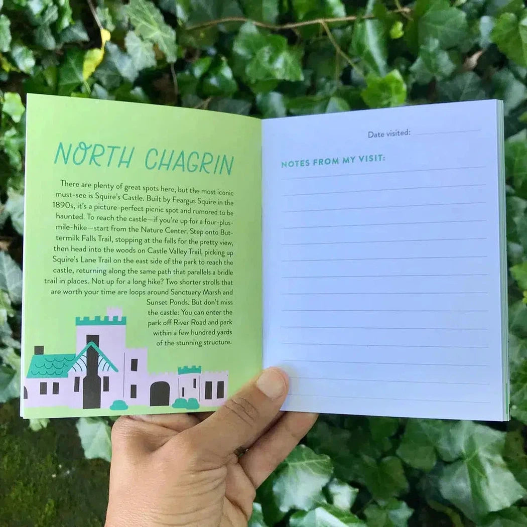 A hand holds open the Free Period Press Cleveland Metroparks Mini-Guide against green foliage, showing text about North Chagrin and a notes page.