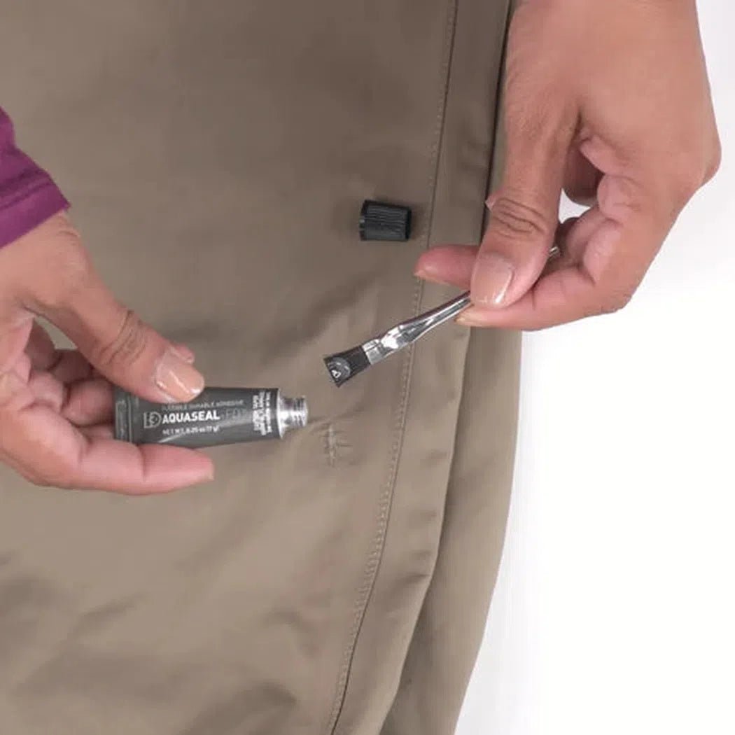 A person holds a tube of Gear Aid Aquaseal FD Repair Kit with a brush applicator, showing the product against a beige background.
