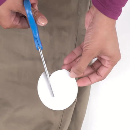 A person uses blue scissors to cut a white circle with the Gear Aid Aquaseal FD Repair Kit against a brown background.