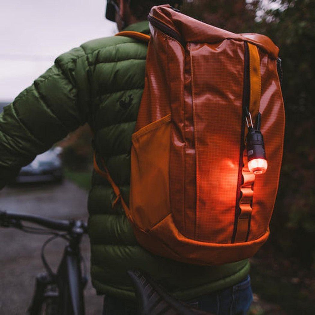 Back view of the Gear Aid Carabiner Light Kit on an orange backpack with a lit light attached, worn by a cyclist in a green jacket.