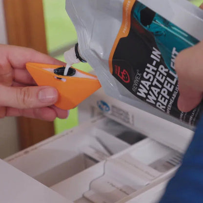 A hand pours Gear Aid Revivex Wash-In Water Repellent into an orange cleaning tool over a white drawer.
