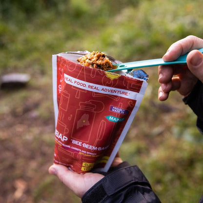 A hand holds a red Good To-Go Bibimbap - Double Serving bag while using a blue spoon to eat from it, with a grassy outdoor background.