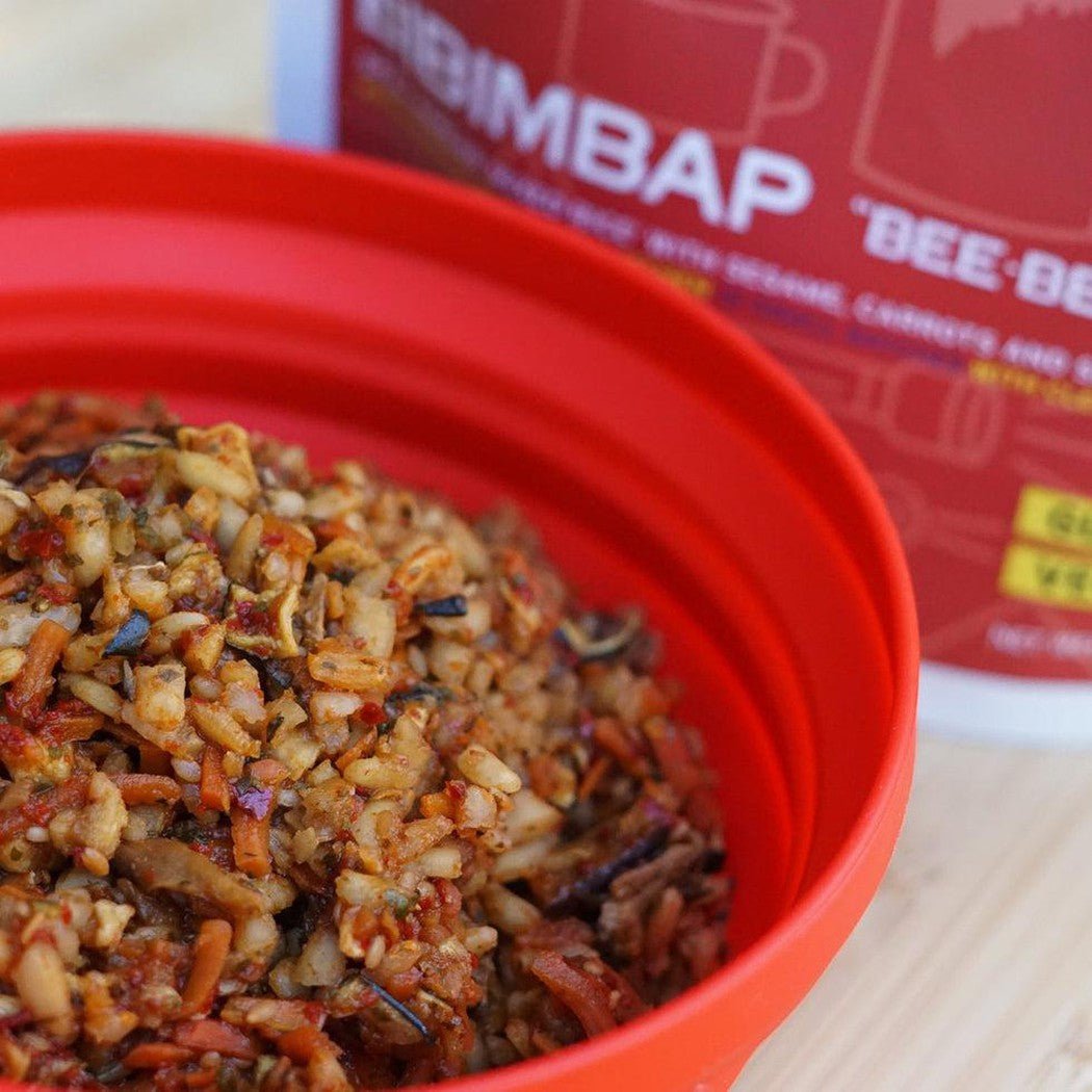 A red collapsible bowl filled with bibimbap sits in front of a Good To-Go Bibimbap - Double Serving container.