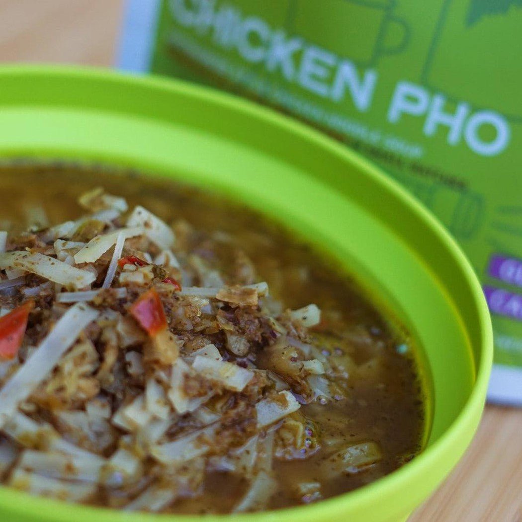 A close-up of the Good To-Go Chicken Pho in a bright green bowl, with the product box visible in the background.