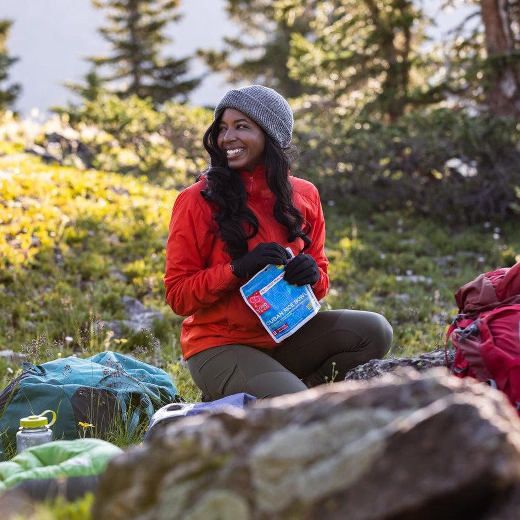 A woman in a red jacket sits outdoors with a blue Good To-Go Cuban Rice Bowl, surrounded by backpacking gear and greenery.
