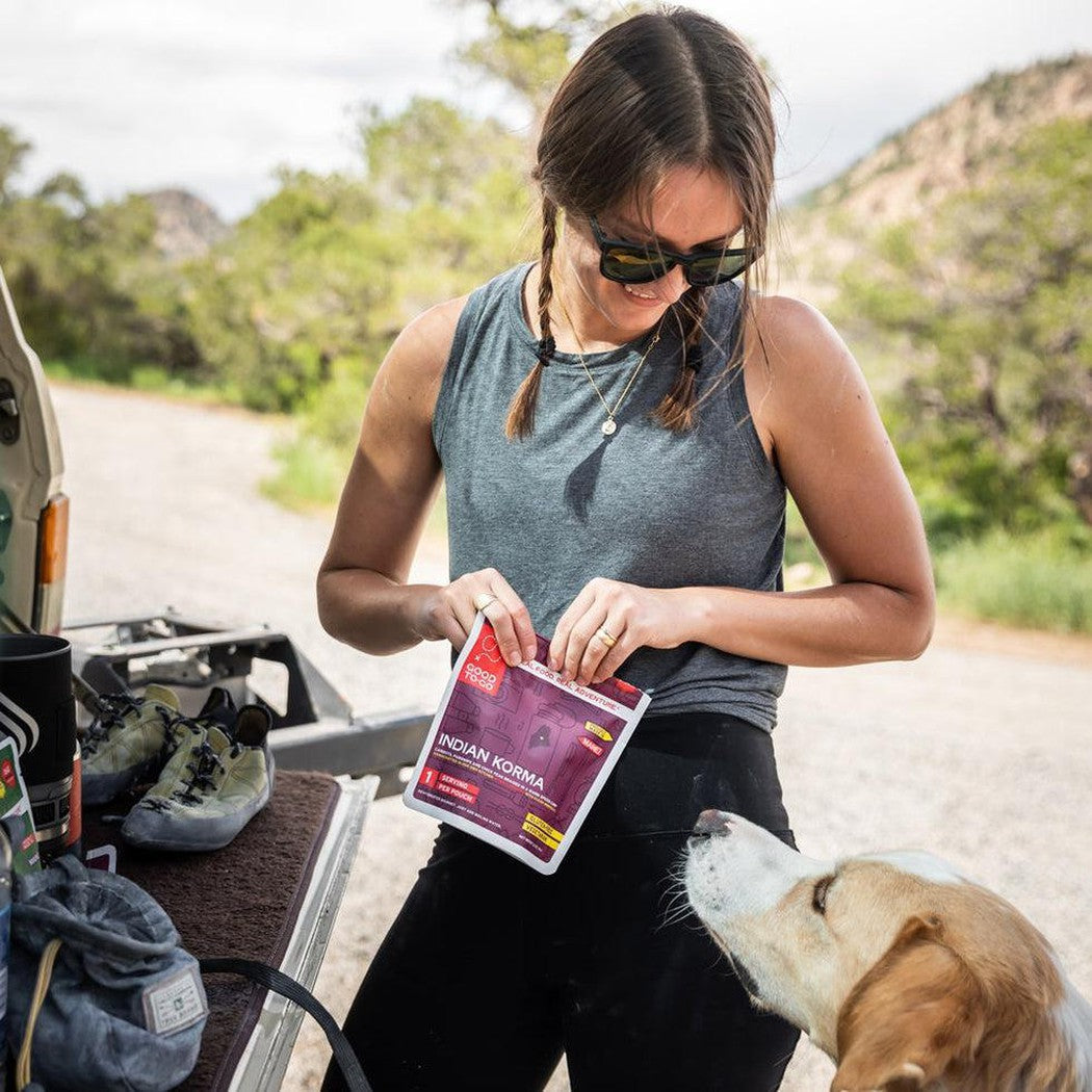 A woman outdoors with her dog holds the Good To-Go Indian Vegetable Korma - Double Serving package, with hiking gear and a scenic road in the background.