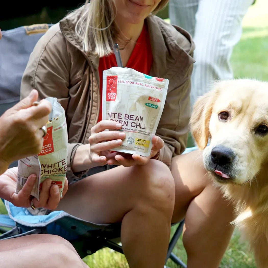 A woman holds a Good To-Go White Bean Chicken Chili pouch while sitting outdoors with a golden retriever nearby.