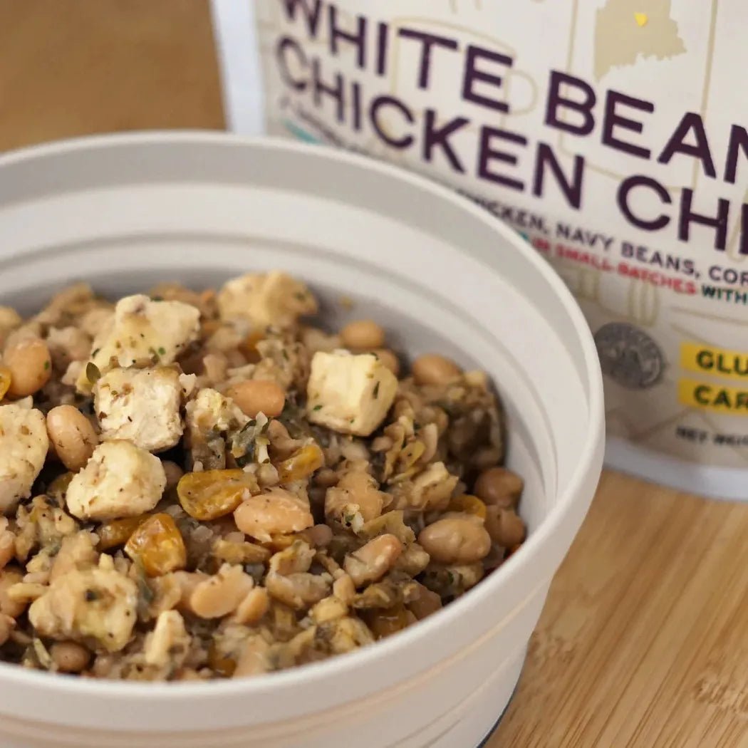 A bowl of Good To-Go White Bean Chicken Chili sits on a wooden surface with the product box in the background.