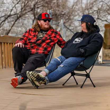 Two people relax in Grand Trunk Caddis HD Chairs on a wooden deck with trees in the background.