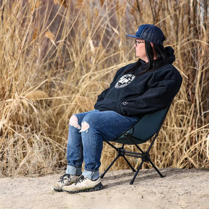 A woman sits in a Storm Blue Grand Trunk Caddis HD Chair on a sandy surface with tall grass in the background.