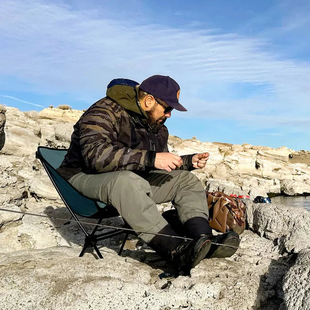 A man sits in a Storm Blue Grand Trunk Caddis HD Chair on rocky terrain near water.