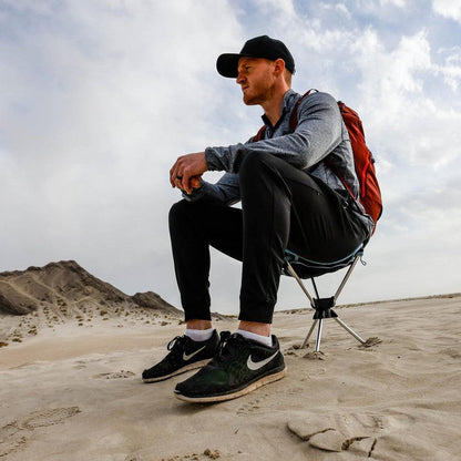 A man sits in a Black Grand Trunk Compass 360 Stool on a sandy desert landscape.