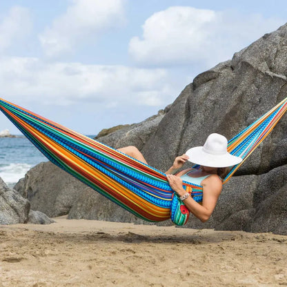 A woman relaxes in a Grand Trunk Double Deluxe Hammock with Suspension Straps on a sandy beach with large rocks and the ocean in the background.