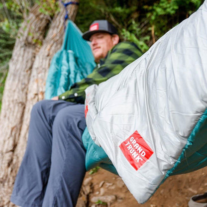 A man in a green plaid shirt sits in a Grand Trunk Evolution Synthetic Hammock, with a silver and teal sleeping bag in the foreground, against a tree and forest background.