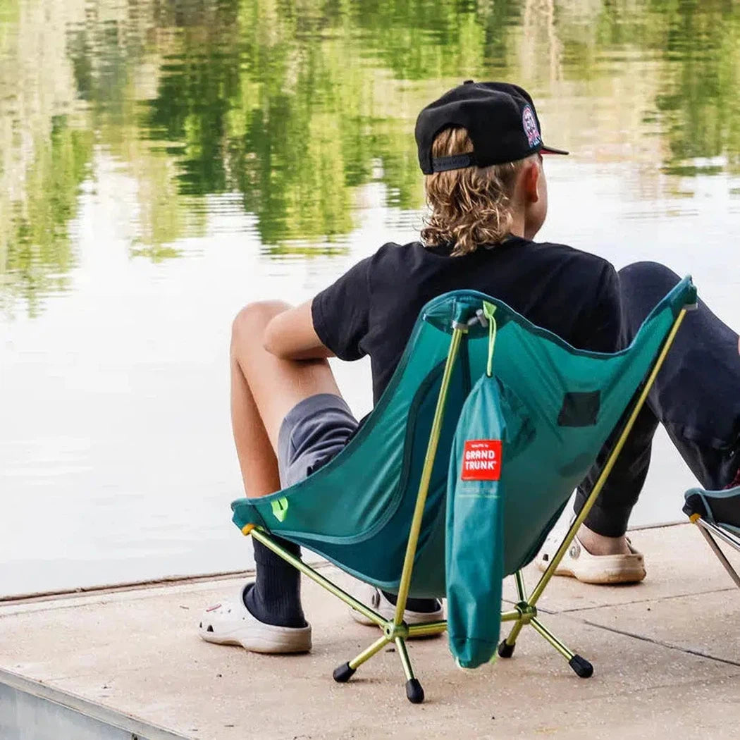 A man in a black shirt and cap sits in a black Grand Trunk Mantis chair by a calm lake.